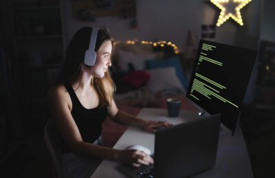 Young girl with headphones and computer sitting indoors, playing games and hacker concept.